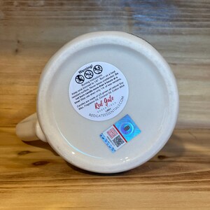 Peut inclure: Gros plan sur le fond d'une tasse en c&eacute;ramique cr&egrave;me avec une anse. Une &eacute;tiquette d'avertissement avec des instructions de combustion et un autocollant holographique sont visibles. Le texte "Red Gate Essentials" est pr&eacute;sent.