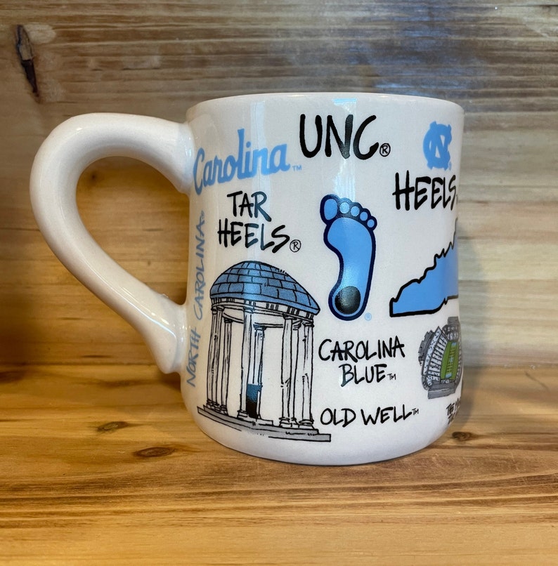Peut inclure: Mug en c&eacute;ramique blanche avec une grande anse, orn&eacute; de divers graphismes et textes bleus et noirs li&eacute;s &agrave; l'Universit&eacute; de Caroline du Nord. La tasse comprend les mots "Carolina", "UNC" et "Tar Heels".
