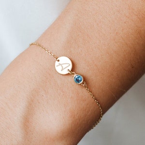 Könnte beinhalten: Ein goldenes Kettenarmband mit einem kleinen runden Anhänger, der mit dem Buchstaben "A" graviert ist, und einem blauen Edelstein.