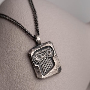 Op de afbeelding: Een zilverkleurige hangerketting met een rechthoekige hanger met een reliëf Grieks kolomontwerp. De ketting heeft een donkere ketting. De ketting wordt gepresenteerd op een neutrale ondergrond.