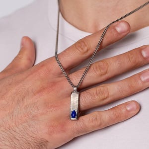 May include: Silver-tone pendant necklace with a rectangular pendant featuring a blue stone inlay. The necklace is worn with a white t-shirt and a black leather bracelet. The pendant hangs on a delicate chain.
