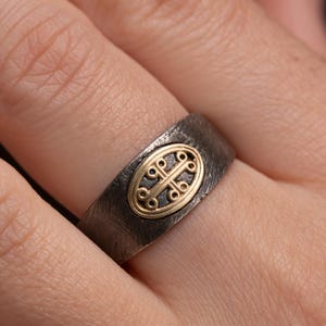 Puede incluir: Un anillo gris oscuro con una superficie texturizada. El anillo presenta una pieza central ovalada de color dorado con un diseño intrincado. El anillo se lleva en un dedo, mostrando los detalles y la artesanía de la joya.