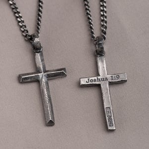 Puede incluir: Dos collares con cruces plateadas y cadenas de eslabones. Una cruz tiene el texto "Joshua 1:9" grabado. Las cruces se muestran sobre una superficie de tono neutro. Los collares son probablemente de metal.