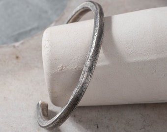 May include: A silver bangle bracelet with a textured finish. The bracelet is open and has a simple, minimalist design.