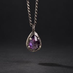 May include: A dark silver-toned chain necklace featuring a teardrop-shaped pendant with a faceted purple gemstone set in a textured metal frame. The pendant hangs vertically from a twisted link chain, showcasing a deep violet hue and metallic finish.