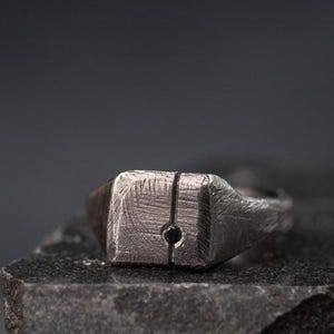 以下が含まれることがあります： 正方形の顔と中央に小さな黒い点がある銀のシグネットリングです。