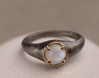 May include: A silver ring with a round, iridescent moonstone set in a gold bezel. The ring has a dark, textured band and a polished gold setting around the gemstone. The ring is displayed on a white surface.