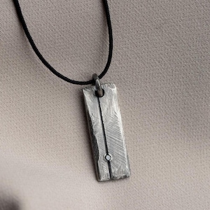 May include: A rectangular silver-toned metal pendant with a textured, brushed surface and a vertical line detail featuring a small circular accent. The pendant hangs from a thin, smooth black cord, creating a simple and clean jewelry piece.