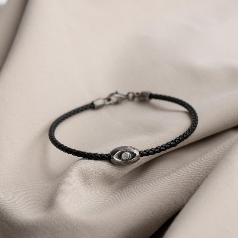 A men's bracelet featuring black leather and an oxidized sterling silver evil eye charm in a brutalist style, perfect for a bold, unique gift.