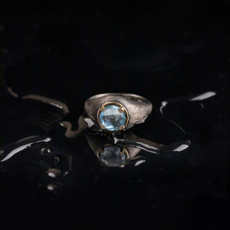 Rustic wide ring in oxidized sterling silver with a handmade 14K yellow gold bezel and a natural blue topaz gemstone on it.