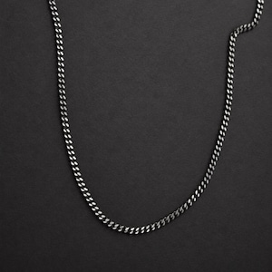 Può includere: Una collana a catena gourmette in metallo grigio canna di fucile scuro con maglie ovali intrecciate. La catena metallica presenta una finitura lucida che riflette la luce lungo i bordi curvi di ogni maglia, creando un filo continuo e flessibile.