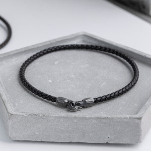 Könnte beinhalten: Ein schwarzes Lederarmband mit einem silbernen Verschluss. Das Armband ist geflochten und hat ein schlichtes Design.