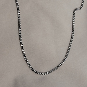 Può includere: Una collana a catena curb argentata su uno sfondo di tessuto marrone chiaro.