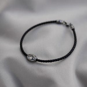 Men's bracelet with a sleek black leather band and a rugged, oxidized sterling silver evil eye, combining contemporary style with brutalist influence.