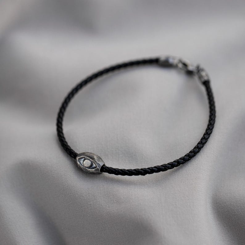 Men's bracelet with a sleek black leather band and a rugged, oxidized sterling silver evil eye, combining contemporary style with brutalist influence.