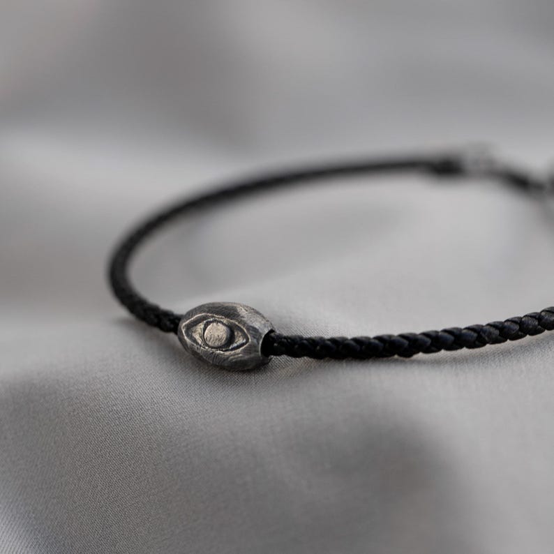 Modern men's bracelet with black leather strap and oxidized sterling silver evil eye symbol in a raw, brutalist design
