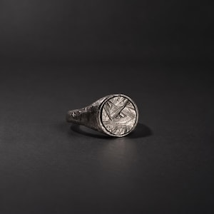 May include: A silver signet ring featuring a circular face with a deeply etched, multidirectional linear pattern. The band and the setting have a dark, oxidized finish with a rugged, uneven texture throughout the metal surface.