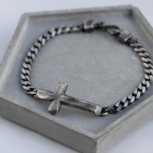 May include: A silver-toned bracelet with a cross pendant. The bracelet features a thick curb chain design and a textured cross. The bracelet is displayed on a gray, hexagonal tray.
