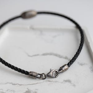 A men's bracelet featuring a strong black leather band and oxidized sterling silver evil eye element, with a modern brutalist lobster clasp.
