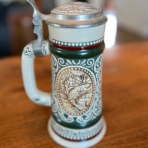 Pode incluir: Uma caneca de cerveja de cerâmica com tampa de estanho, apresentando uma ilustração detalhada de um peixe em uma moldura oval verde e marrom. A caneca tem um fundo branco com detalhes verdes e vermelhos.