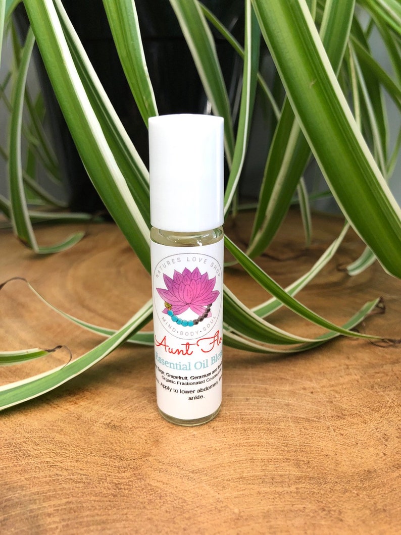 May include: A white roller bottle with a pink lotus flower and the text "NATURES LOVE SHOP MIND BODY SOUL Aunt Flow Essential Oil Blend Grapefruit, Geranium and Organic Fractionated Coconut Oil Apply to lower abdomen or ankle."