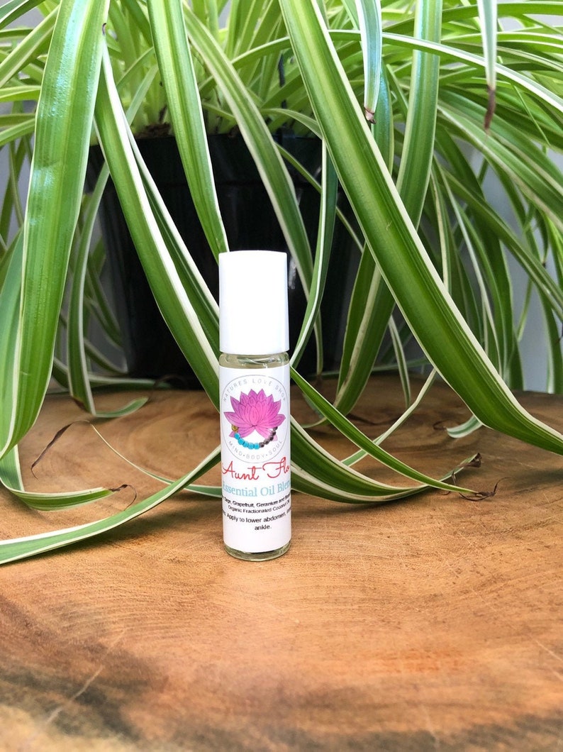 May include: A clear glass roller bottle with a white label. The label has a pink lotus flower and the text "Aunt Flow Essential Oil Blend" with a list of ingredients.