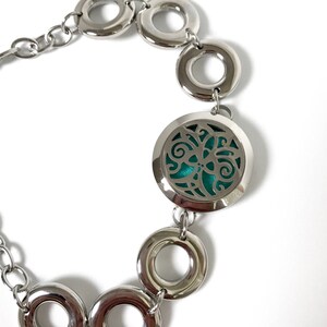 May include: A silver bracelet with a round, open locket featuring a teal-coloured, floral design. The bracelet is made of a chain with multiple silver rings.