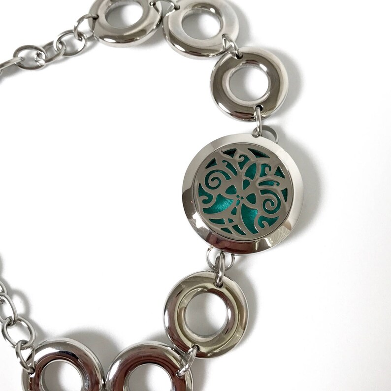 May include: A silver bracelet with a round, open locket featuring a teal-coloured, floral design. The bracelet is made of a chain with multiple silver rings.