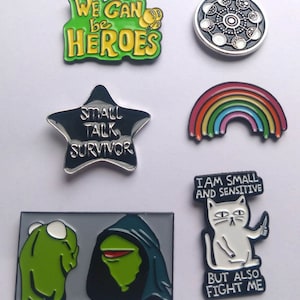 May include: Set of 7 enamel pins featuring various designs.  The pins include a green and yellow pin with the text "WE CAN be HEROES", a black and silver star-shaped pin with the text "SMALL TALK SURVIVOR", a rainbow-coloured pin shaped like a rainbow, a silver pin with a circular design, a black and white pin with a cat holding a knife and the text "I AM SMALL AND SENSITIVE BUT ALSO FIGHT ME", and a green and black pin featuring a meme of Kermit the Frog in a hooded robe.