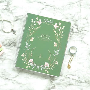 May include: A green 2027 Catholic planner with a white spiral binding, featuring a floral wreath illustration on the cover. The cover includes the text 2027, Catholic Planner, and a small gold emblem. Accessories include a white beaded rosary, three pastel rectangular markers, a gold paperclip with a religious charm, and a small roll of tape.