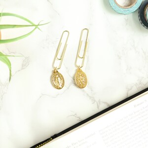 May include: Two gold paper clips with gold charms attached. The charms are oval shaped and feature a religious design. The paper clips are on a white marble surface.