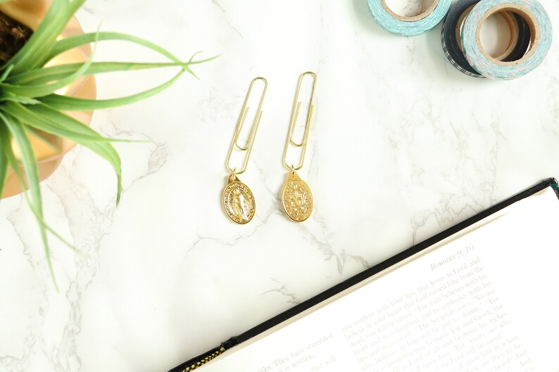May include: Two gold paper clips with gold charms attached. The charms are oval shaped and feature a religious design. The paper clips are on a white marble surface.