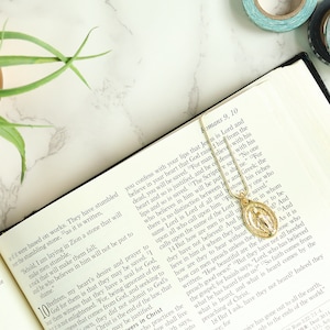 May include: An open book with a gold chain and pendant necklace with a religious image. The book is open to a page with text from the Bible, Romans 9:10. The book is on a white marble surface with a green plant in a gold pot.