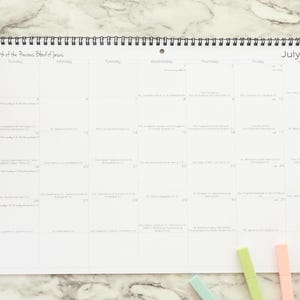 May include: A white, spiral-bound calendar for July 2026, with a grid layout and handwritten notes. The calendar is on a marble surface, with colorful markers and a small plant in the corner.