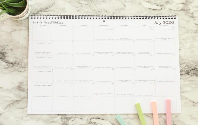 May include: A white, spiral-bound calendar for July 2026, with a grid layout and handwritten notes. The calendar is on a marble surface, with colorful markers and a small plant in the corner.