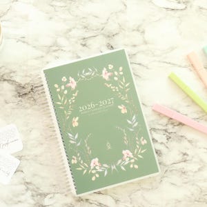 May include: A green 2026-2027 planner with floral accents and a spiral binding. The planner is surrounded by pastel-colored pens, decorative tape, and cards. A small vase of red flowers is in the upper left corner.