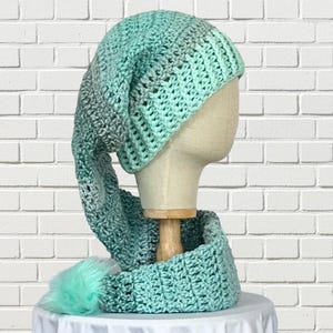 May include: A handmade crochet hat and scarf set in shades of teal, gray, and white. The hat has a long, pointed shape, and the scarf is a loop style. A teal pom-pom adorns the end of the scarf. The set is displayed on a mannequin head.