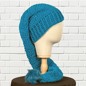 May include: A teal blue, handmade crochet hat with a long, pointed tail and a fluffy pom-pom. The hat is displayed on a mannequin head against a white brick wall. The hat is made of a thick, textured yarn.