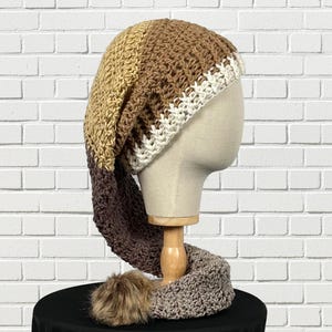 May include: A handmade crochet hat and scarf set displayed on a mannequin head. The hat features a long, pointed design with color blocks of tan, brown, and white. The matching scarf is gray with a brown faux fur pom-pom.
