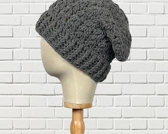 May include: A gray, crocheted beanie hat displayed on a mannequin head. The hat is made of a thick, textured yarn and has a slouchy fit. The mannequin is on a wooden stand, set against a white brick wall.