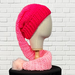 May include: A handmade, crochet hat and scarf set in shades of pink and red. The hat is a vibrant fuchsia, with a long, pointed end. The scarf is a gradient of pinks, and the set includes a fluffy, light pink pom-pom.