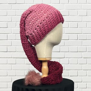 May include: A handmade, crochet hat and scarf set in shades of pink and burgundy. The hat has a long, pointed end, and the scarf features a fluffy, pink pom-pom. The set is displayed on a mannequin head against a white brick wall.