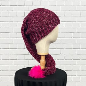 May include: A long, deep burgundy knit hat with a slouchy, pointed design that extends downward. The hat features a thick, textured yarn construction and is finished with a bright, vibrant hot pink pom-pom at the very tip of the tail.
