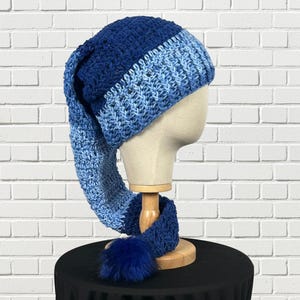 May include: A long, slouchy crochet hat featuring a gradient of blue yarn, transitioning from a deep navy at the top to a lighter sky blue at the base. The hat includes a ribbed brim and a dark blue faux fur pom-pom attached to the end of the tail.