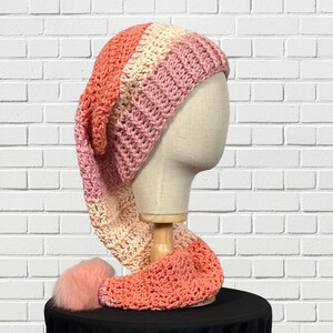 May include: A handmade, long, crochet hat in a gradient of colors, including peach, pink, and cream. The hat has a long, pointed end and a matching scarf-like extension. A fluffy, peach-colored pom-pom adorns the end of the scarf.