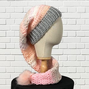 May include: A handmade crochet hat and scarf set in shades of pink, peach, and gray. The hat has a long, pointed shape and a gray top. The scarf is attached and features a pink pom-pom at the end. The set is displayed on a mannequin head.