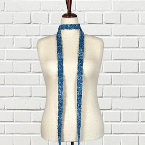 May include: A blue knitted scarf displayed on a mannequin. The scarf is a long, narrow design with a textured appearance. The color is a vibrant blue with lighter blue accents. The scarf is displayed against a white brick wall.