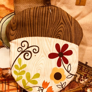 May include: A brown and white acorn-shaped pot holder with a wood grain pattern and floral designs in shades of red, yellow, and green. The pot holder has a loop for hanging. The text "PARK Designs" is visible on a brown tag.