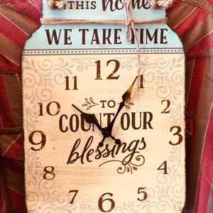 Puede incluir: Un reloj de color azul claro en forma de tarro de albañil con fondo blanco y números marrones. La esfera del reloj dice "En este hogar nos tomamos el tiempo para contar nuestras bendiciones".