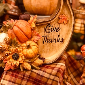 以下が含まれることがあります： 三日月型の木製看板に「Give Thanks」の文字が書かれています。看板は、小さなカボチャ、ひまわり、トウモロコシの穂など、秋のモチーフで飾られています。看板は格子模様のテーブルクロスの上に置かれています。